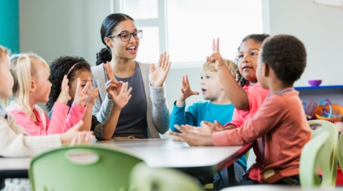 Teacher sits at a table making a clapping motion with diverse preschool class