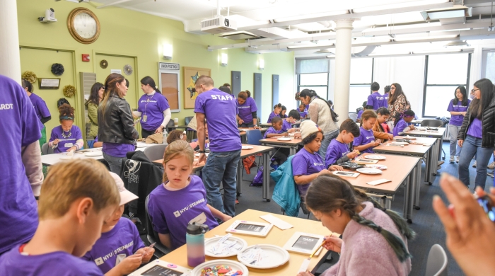 A crowd of volunteers and children complete a craft in an art classroom.