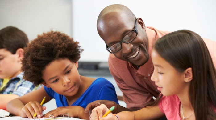 Educator assists two elementary school students as they sit at their desk completing class assignment. An Blac female is seated to the left of the teacher, while a Latina students sits to the right of the male teacher wearing Black frame eye glasses.