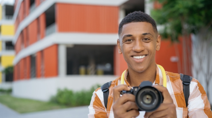 Captures image of student photographer holding his camera, as he smiles while his own photo is being taken.