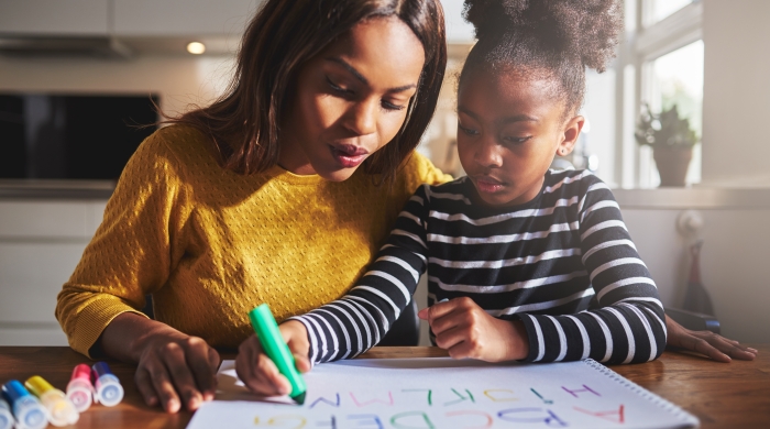 Image captures a Mother assisting her daughter, as she writes out her alphabet on a large sheet of white paper.
