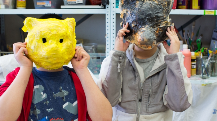 Two children hold paper mache animal masks to their faces.