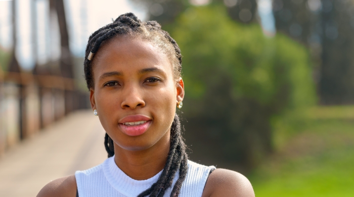 Image captures an African American student hailing from rural community, sitting in front of bush in a green field adjacent to a dirt road.