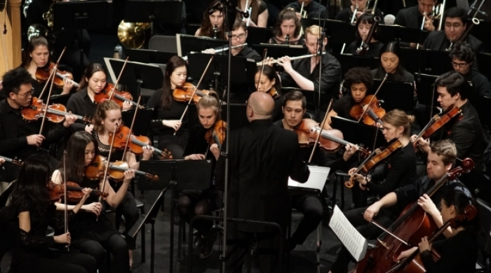 A conductor stands on the podium surrounded by NYU student musicians.