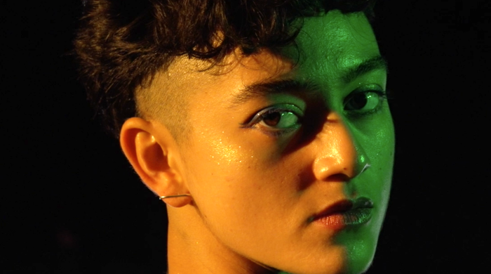 Image of Luna Beller-Tadiar, a queer person with short black hair, shaved sides, and light brown skin. She is lit by green and orange light and looks at the camera.