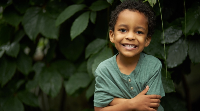 Image captures a young child in a green pasture. The elementary school student stands directly in front a full growth of thick leaves.The little Black boy smiles with his arms folded. The child wears a green colored t-shirt, that is nearly the exact same color as the leaves behind him.color