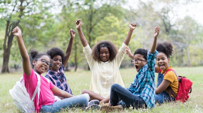 Image captures 4 Black students and their teacher sitting in a park. Each member of the groups sits on green grass, amongst a field of tall green trees. The 4 students are sitting in a circle around the teacher wearing a white colored sweater. Each student has a backpack on their back, as each throughs both of hands in the air, along with their teacher/