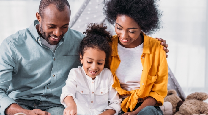 Image captures a family reading a book together. Two parents are seated on the floor with their elementary school aged child between them. Both the mother and father assist the child in reading  a book. The father wears blue shirt, while the mother wears a yellow sweater.