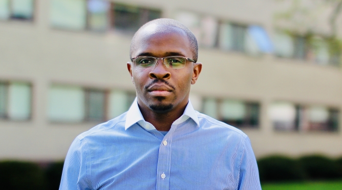 A black man wearing a blue oxford shirt and glasses looks at the camera