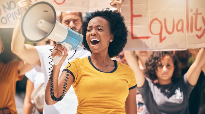 Image captures Woman protesting with a group of people. The is group marches in the street, as the photo centers around a woman yelling into an electric bullhorn. The Woman wearing a yellow shirt, and holds a white and blue bullhorn in her right hand.