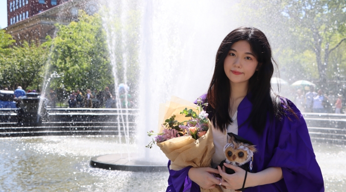 A yoing asian woman in graduation garb stands in front of a public fountain holding flowers. 