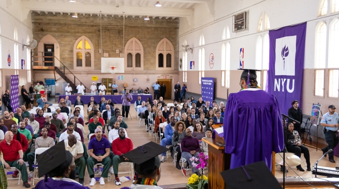 Overlooking an audience, a man wearing a violet graduation robe and cap gives a speech