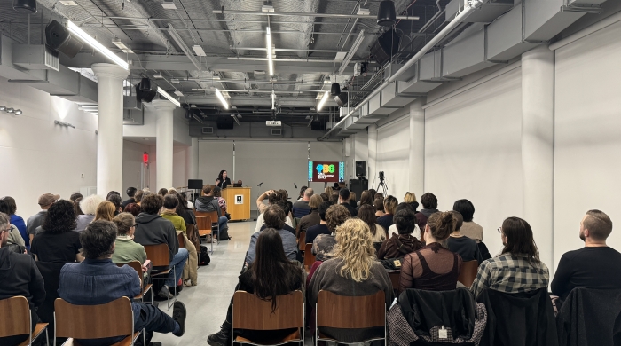 A large auditorium listens to a lecture delivered by someone at a podium. A screen displays the original logo for Publlic Broadcasting.