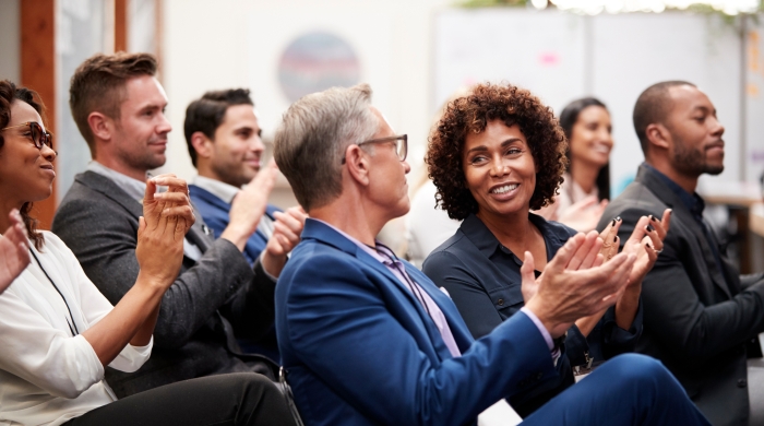 Image captures the a seated conference audience. The photo centers around two audiences members sitting together.. Nearest the camera lens, is a man wearing a blue suit, as he applauds. Next to him, is Black Woman who is in conversation with the man in the blue suit.