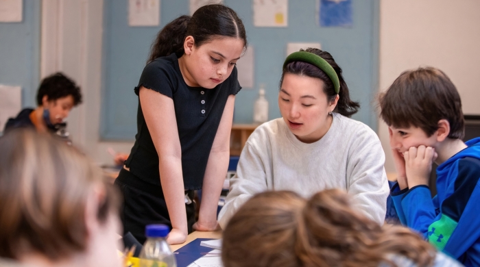 In a classroom, a teacher and student look at something on the desk together