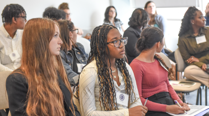 This snapshot captures attendees of NYU Metro Center's 2025 Equity Conference. The attendees make up the audience for breakout sessions.. Th photo captures more thn 12 individual in the breakout session. The image itself is centered around3 figures un the frount row. The figure on the lleft wears a black blazer. She also has long red hair. The figure inthe middle wears a white toop and sports lon black braids. This inidivual also has items in her lap to take notes during the session. Lastly, the indidual t 