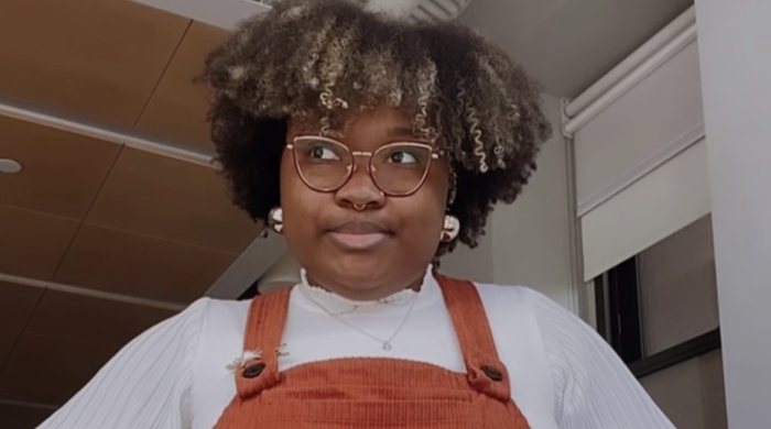 Photo of Imani posing with her hands on her hips, wearing a white long sleeve top and orange overall dress