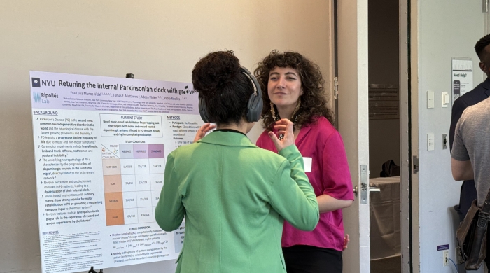 PhD student stands to the left of their poster while an attendee stands in front of them with headphones on listening to an example of the student's rehabilitative research using music.