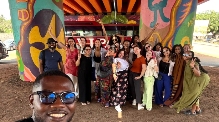 A smiling man in sunglasses takes a selfie while a group of students poses in the background under a brightly-painted bridge