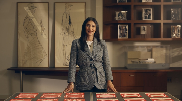 A woman with black hair smiles facing the camera. She is standing behind a table with printouts of red graphics.