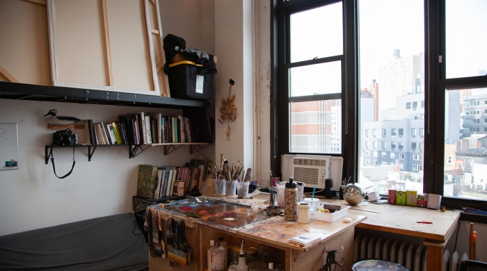 An artist studio featuring a desk center frame with paint brushes and supplies on it, a book case on the left, and large windows looking out into the city on the right.
