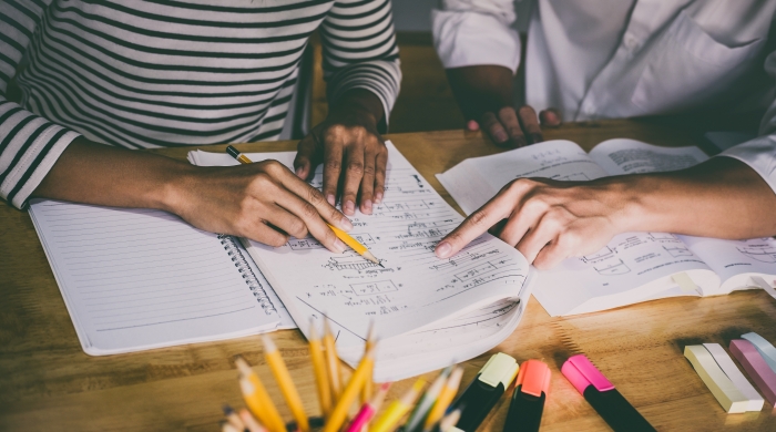 A student's hand holds a pencil to a sheet of paper, while a teacher's hand points at the writing on the page. 