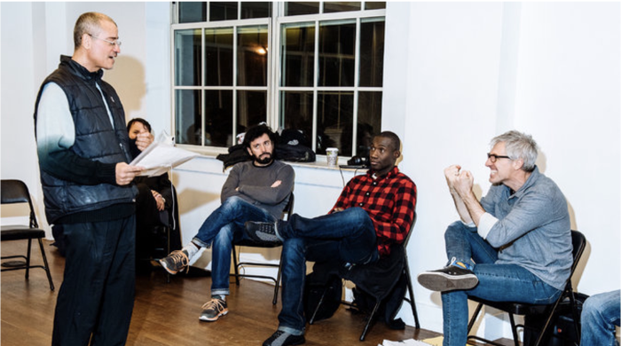 A performer stands, holding a piece of paper, getting feedback from an instructor while a small, seated group observes