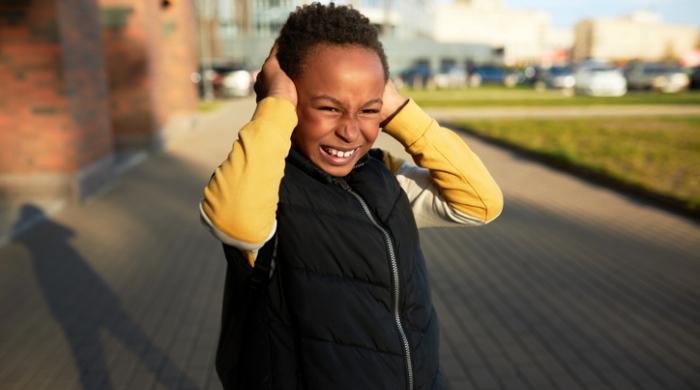 Image captures a child covering his ears. The African-American child wears a Black vest over a yellow long sleeve shirt. The chold stand in the outdoors and raisses his hands to conver his ears, to protect themselves from a lourd peircing noise. 