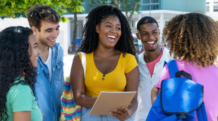 image captures 5 high school students gathered togeter. The students sit in a circle as they wor together. The photo is centerd around an African American female with shoulder lnegth hair, wearing a yellow top.