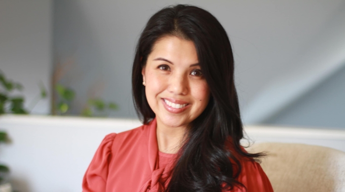 Headshot of Aimee Hilado smiling at the camera, wearing a coral blouse with a tie detail, with long dark hair and a softly lit indoor background with plants.