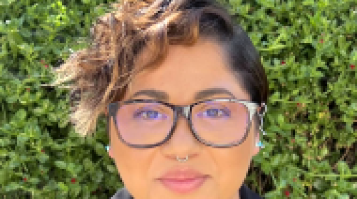 Headshot of Kay Varela smiling slightly at the camera, wearing glasses and a black blazer, standing in front of green foliage outdoors.