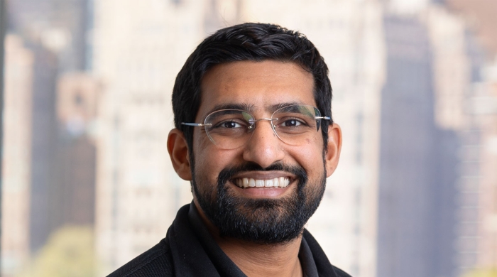 Headshot of Nikhil Garg smiling at the camera, wearing glasses and a dark jacket over a collared shirt, with a softly blurred city skyline in the background.