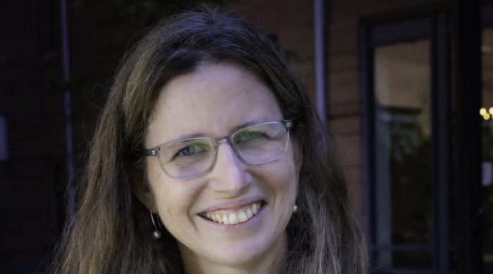 Headshot of Milagros Nores smiling at the camera, wearing glasses, a dark blazer, and a statement necklace, with long light brown hair and an outdoor building background.