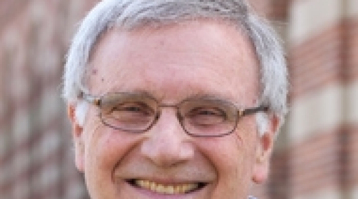 Headshot of Tom Weisner smiling outdoors, wearing glasses and a navy sweater over a light blue collared shirt, with a brick building in the background.