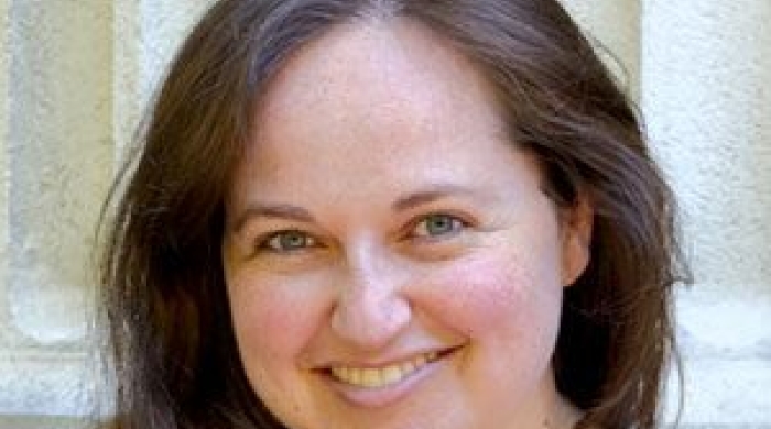 Headshot of Valerie Shapiro smiling at the camera, with long brown hair and wearing a patterned blouse, standing in front of a light stone wall.