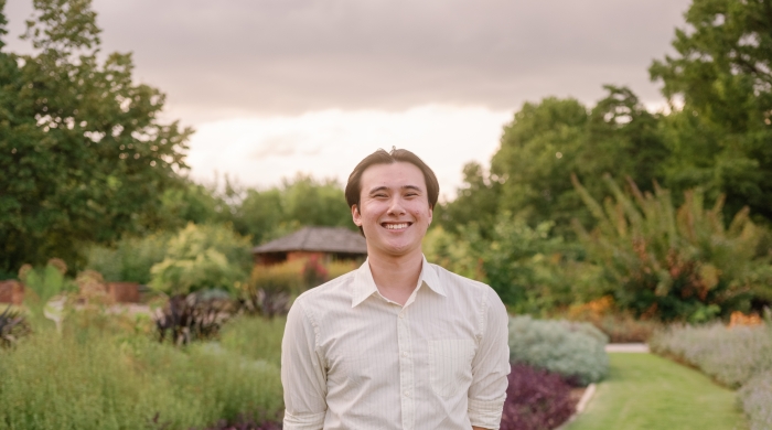 The photo shows a professional photo of Christian Wong in a garden posing for a photo in a white button down shirt and navy blue pants 