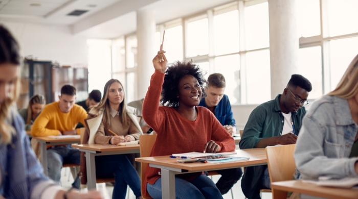 Image captures a school classroom full of studeents. the studets are sitting in rows of individual desks. The Camera focuses on a B lack femalkes studwnt wearing a red sweatshirt. The student smiles ear to ear, as she holds her arm up to confideltly answers a question poosed by her teacher.