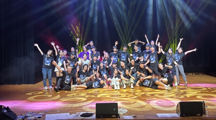 A large group of people pose for the picture on a stage, standing in a group with arms out under bright stage lights