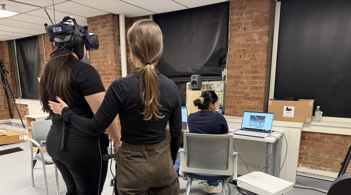 Participant stands on balance board while wearing VR headset, while one research assistant supports them with a hand on their back, and another research assistant operates a computer nearby