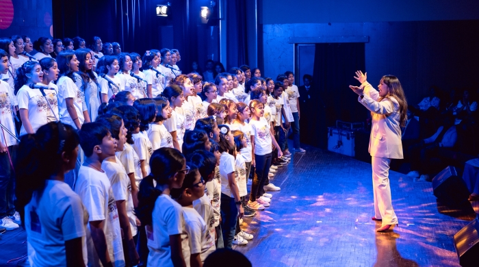A conductor in a white suit leading a large choral group of school age kids