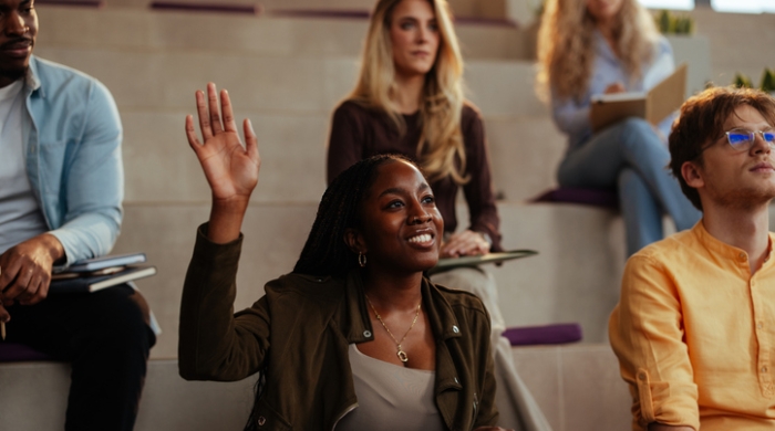 image captures an image of a group of educators attending a professional learrning event. The teachers are seated and listening intently to a speaker. The photo centers around a Black Woman wearing a brown sportcoat. She has her hand raised to engage the moderator. Her hair is breaded and scultpted into a ponytail. She is seated in-between a gentleman wearing a yellow shirt on the right, and a blue top on the left.