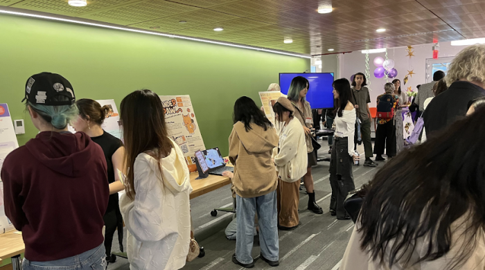 Students navigate around tables with boards and computers showcasing student work. 