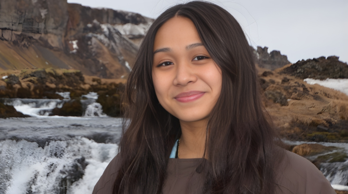 A young woman with long dar brown hair parted in the middle stands in front of a waterfall. She has a camera strap around her neck and is wearing a dark brown jacket. 