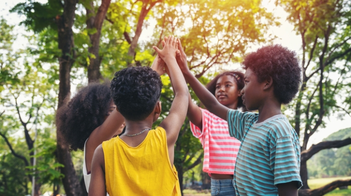 Image captures four students standing in a circle in a green space.. the students collectively raise their hands to reach of the sky,. The student furthest away from the camera wears a pink striped shirt, while the student to the right wears a light blue t-shirt. The student with their back to camera wears a yellow shirt, while the adjacent wears an olive green top.