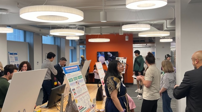 Students stand before research posters at tables in a bustling room. 
