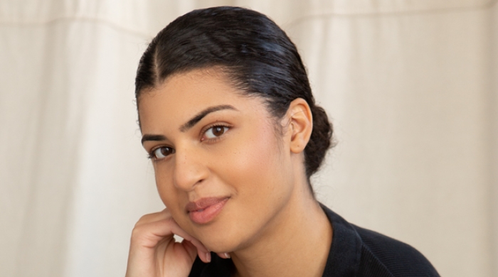 A young woman with tan skin and dark hair sits, looking into the camera.