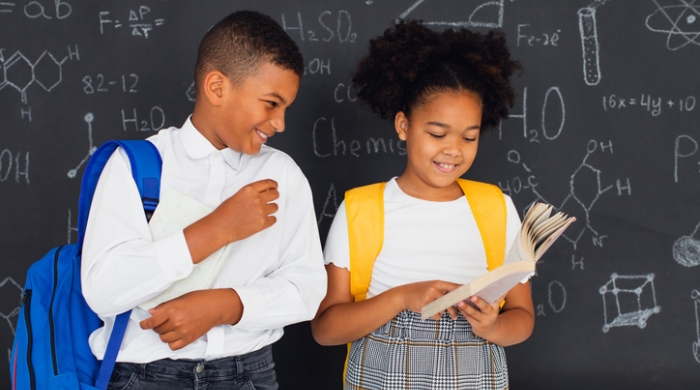 Image captures two students standing in front of chalkboard in their classrooms. One student stands on the left. He wears a white long-sleeve collarded shirt with a blue backpack. On his right, a student wearing a grey skirt and white short-sleeve shirt. and a yellow backpack. 