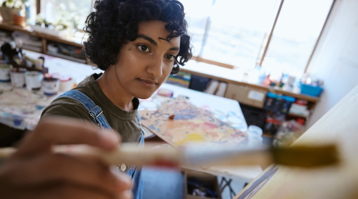 A young artist with curly hair wearing denim overalls focuses on a canvas, holding a paintbrush in the foreground with a wooden palette and sunlit studio in the background.