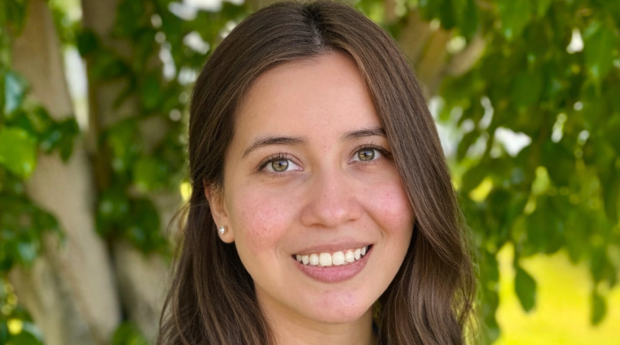 A young woman with brown hair smiles in front of a tree.