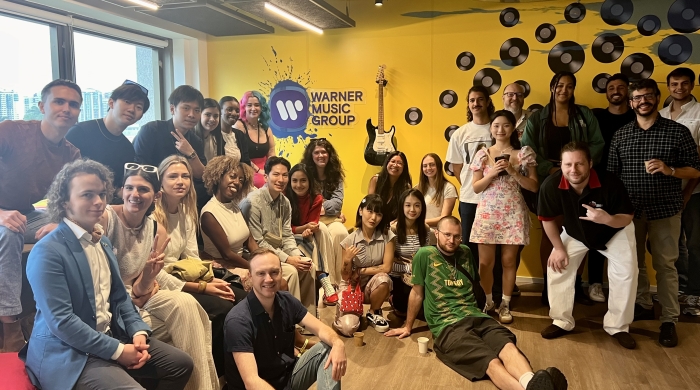 A group of students sits in front of a yellow wall that reads "Warner Music Group"
